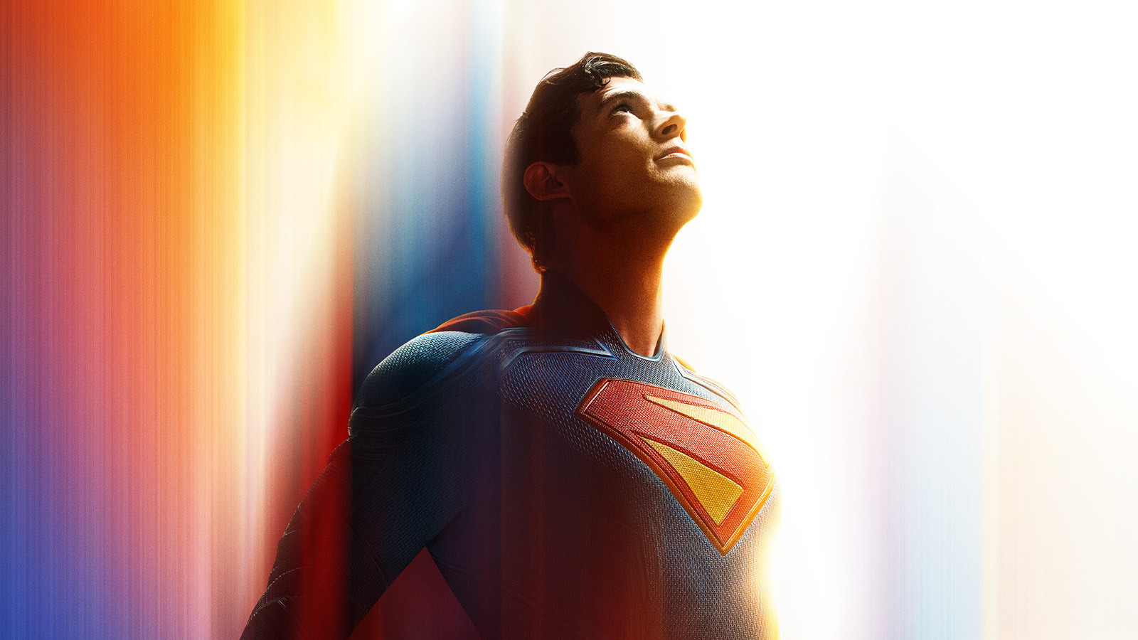 Superman stands and looks at the sky. He is wearing his blue costume with a yellow and red S on the front and his red cape is billowing behind him.