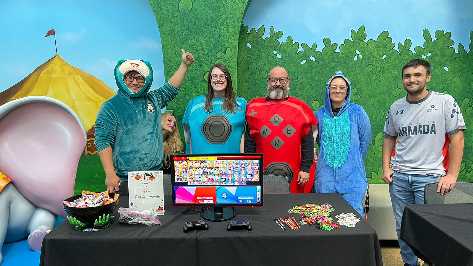 Full Sail staff and students stand next to each other and smile at the camera wearing costumes. A table in front of them has candy, stickers, and a gaming console on it.
