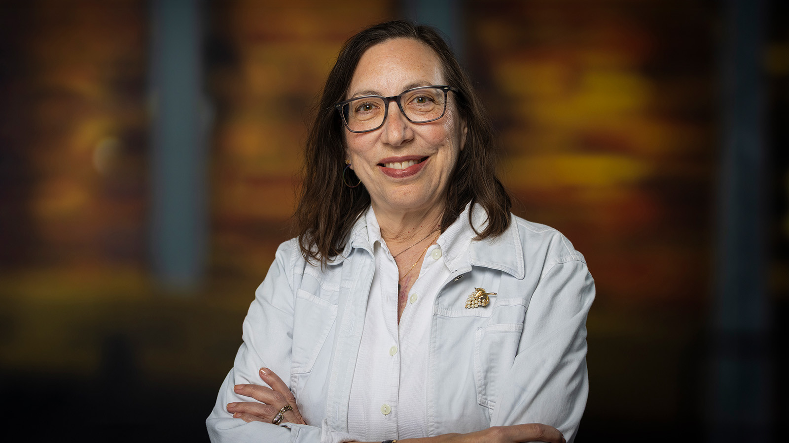 Abby Ginsburg smiles at the camera in front of a wood background. She is wearing a white jacket and glasses.