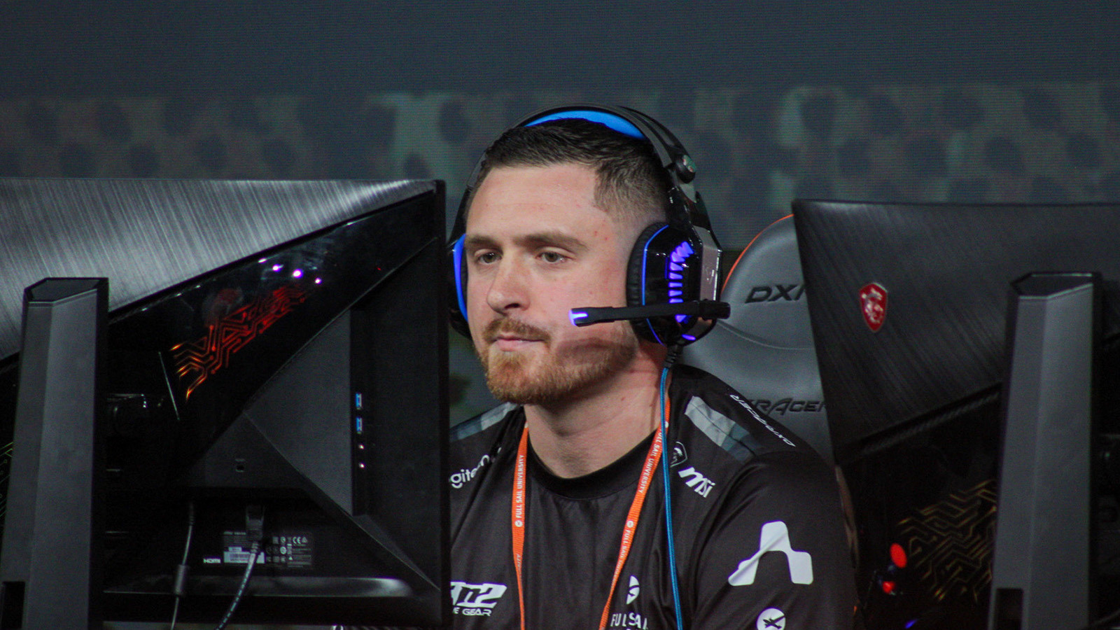 A focused esports player wearing a headset competes at a gaming event, seated between two monitors with a dark, intense atmosphere.