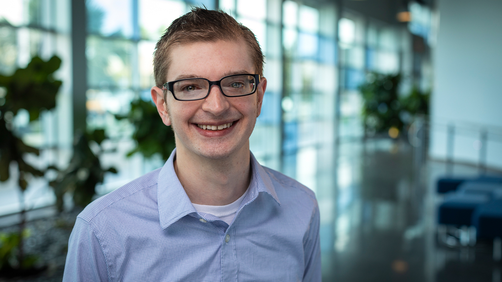 Wes Locher stands in a lobby with lots of windows and smiles at the camera. He wears glasses and a blue checked button-up shirt.