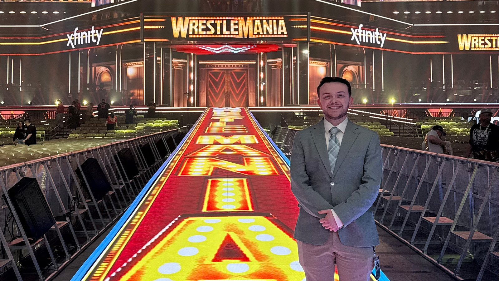Colby Applegate wears a suit and stands in a brightly lit wrestling arena with Wrestlemania graphics displayed on the walls and floor.