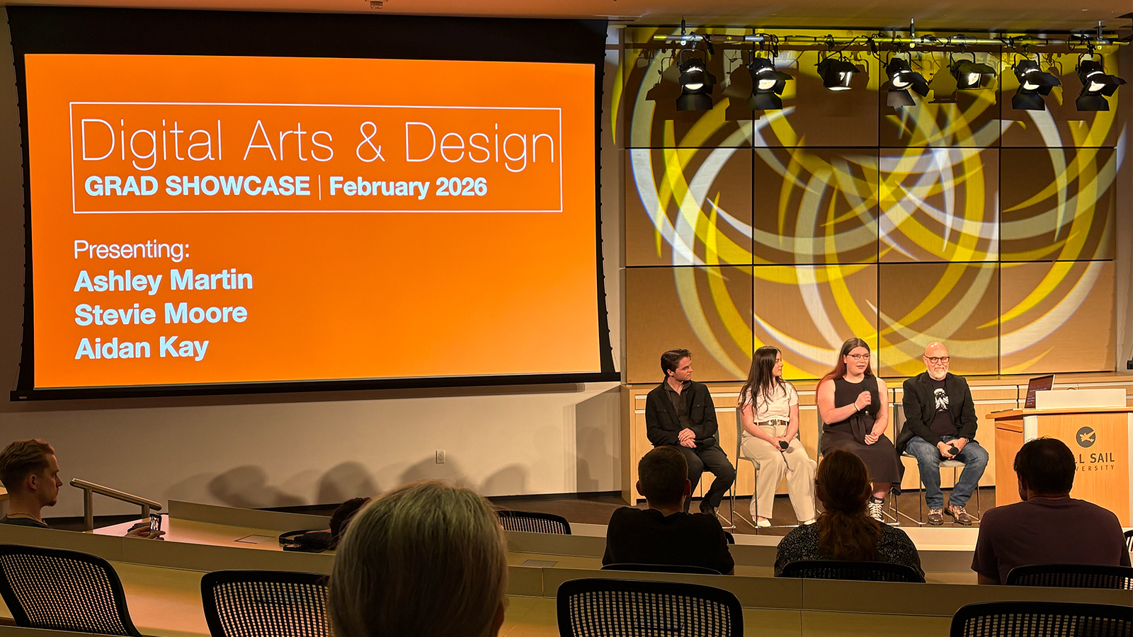 Aidan Kay, Stevie Moore, Ashley Martin, and Joel Sink sit in a row and present to an audience. A large screen behind them displays “Digital Arts & Design Grad Showcase February 2026” and a list of their names.