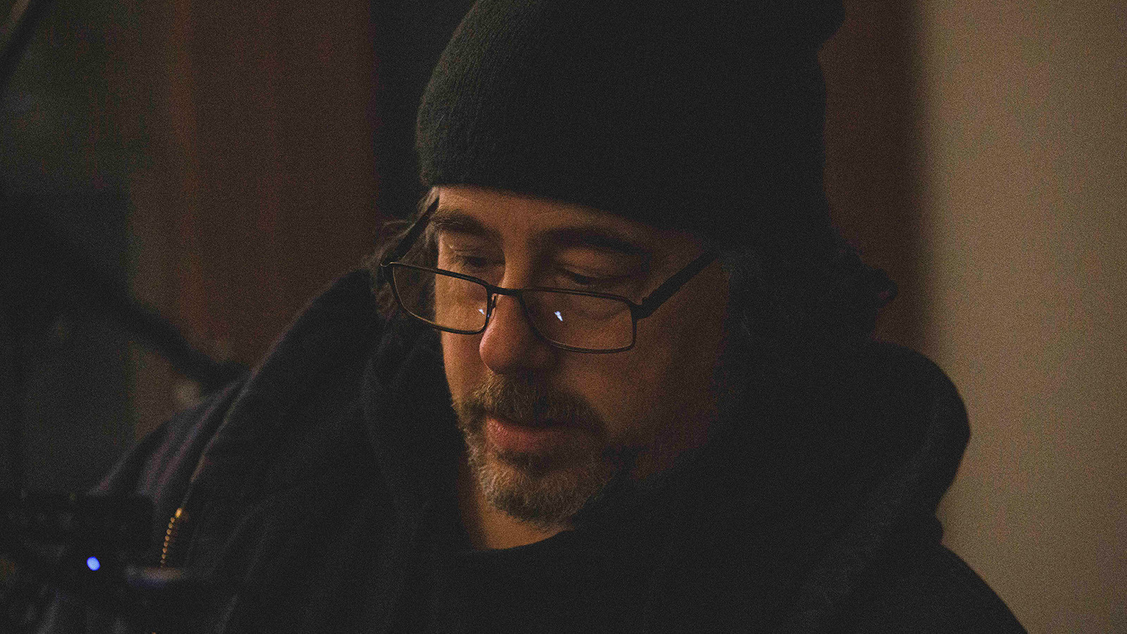 Michael Romano stands in front of a grey wall wearing a beanie, hoodie, and glasses with a pencil tucked behind his ear.
