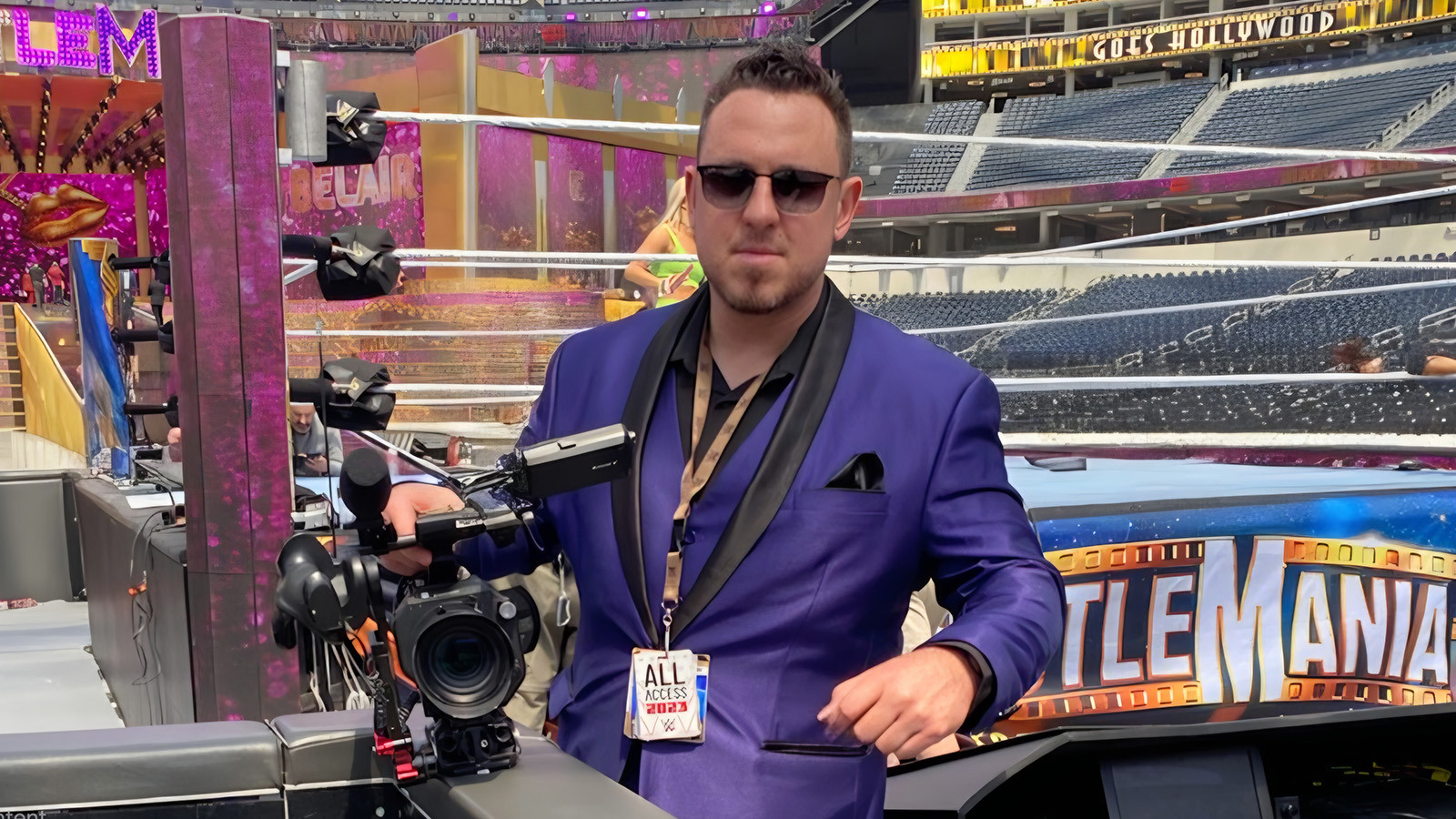 Andrew Karr wears sunglasses and a purple suit as he holds a camera in front of a wrestling ring in a WrestleMania arena.