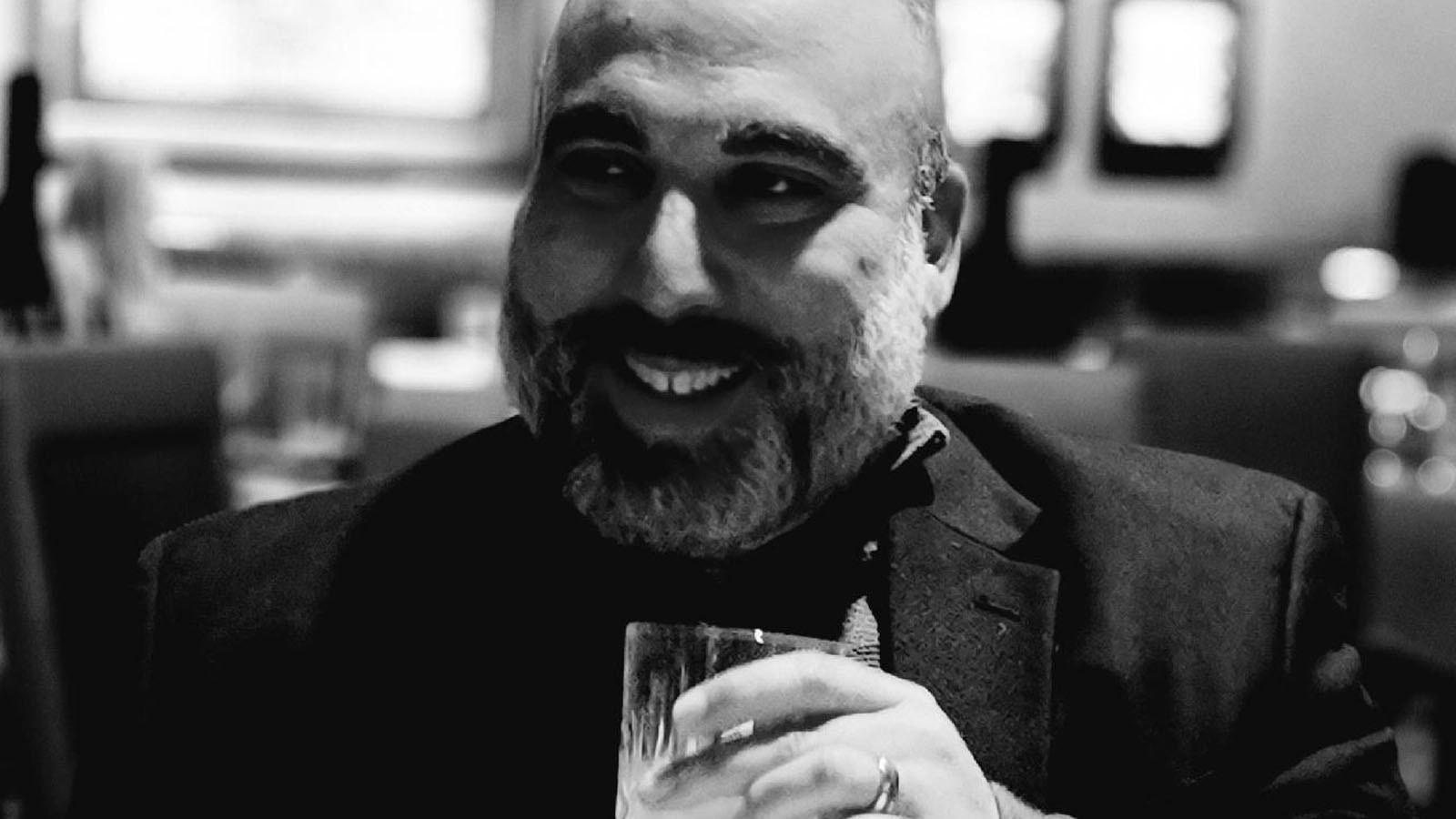 Omar Ortiz smiles as he holds a glass in his hand. He is wearing a dark jacket.