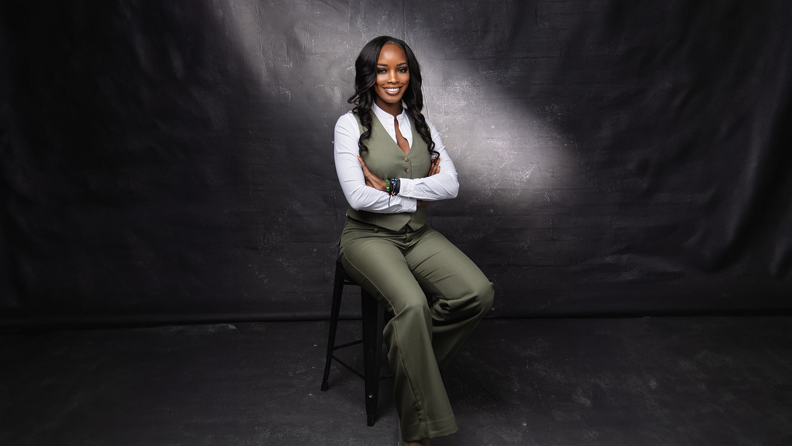 A woman in a green vest and pants sits confidently on a stool, arms crossed, against a dark studio backdrop.