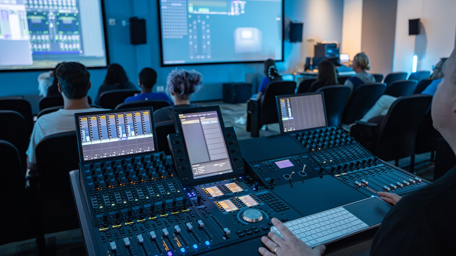 A person is seen operating a professional audio mixing console in a classroom, with students seated and screens displaying sound software.