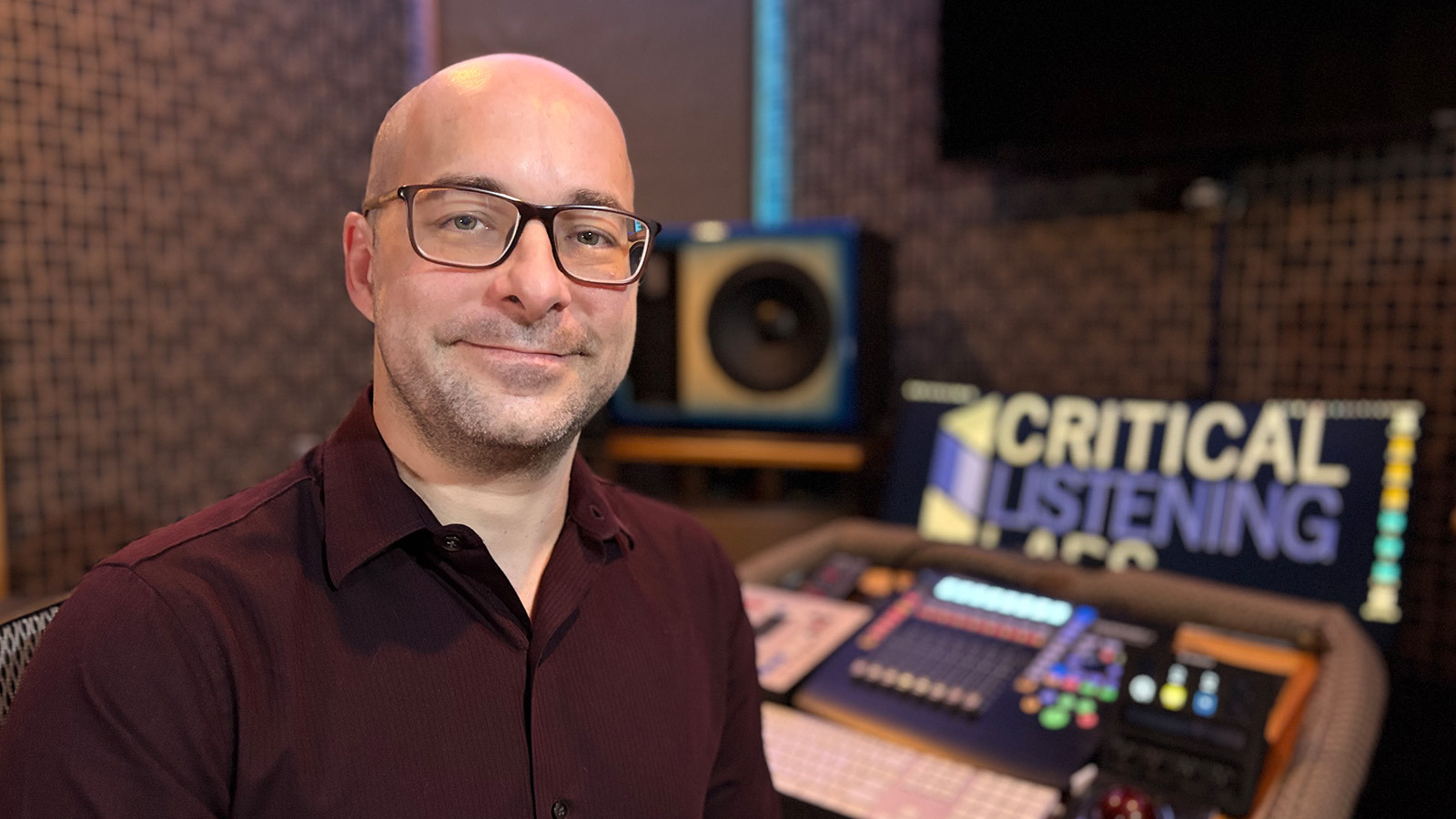 Aaron Gandia sits in a recording studio. He is smiling and wearing glasses and a burgundy shirt. A soundboard is behind him.