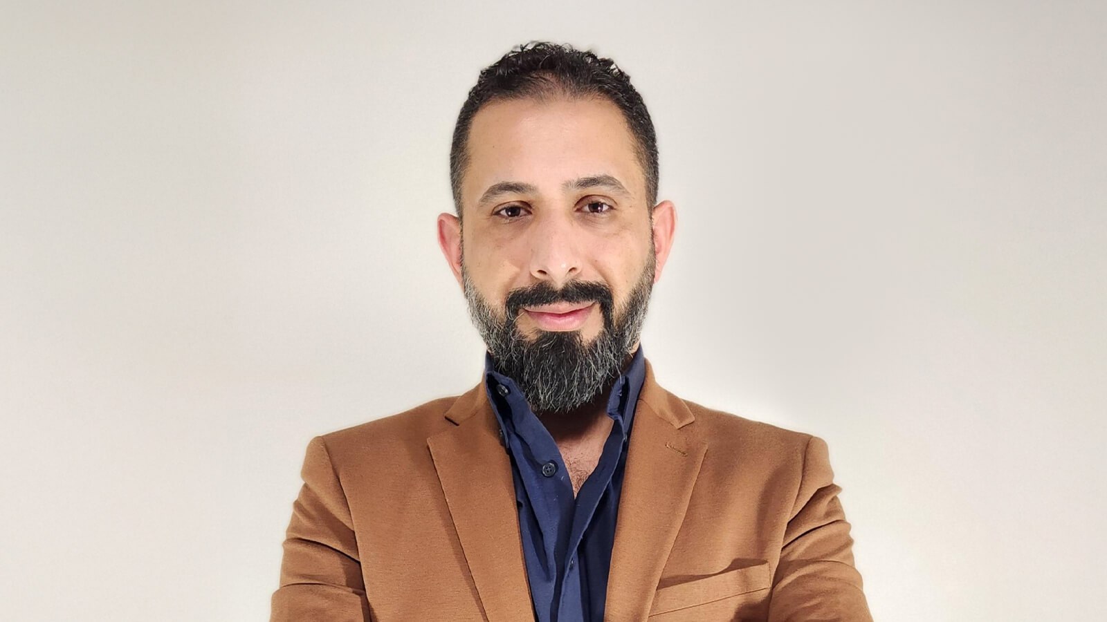 Dr. Ahmed Al Zaidy stands with his arms crossed and smiles at the camera. He is wearing a brown blazer over a blue button-up shirt.