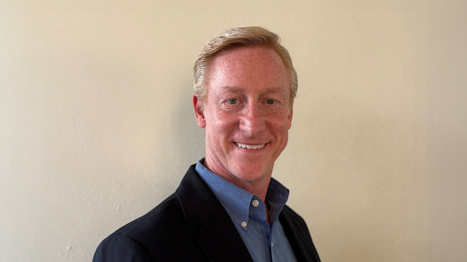 Greg Downey smiles at the camera in front of a light background. He is wearing a blue shirt and a dark blazer.