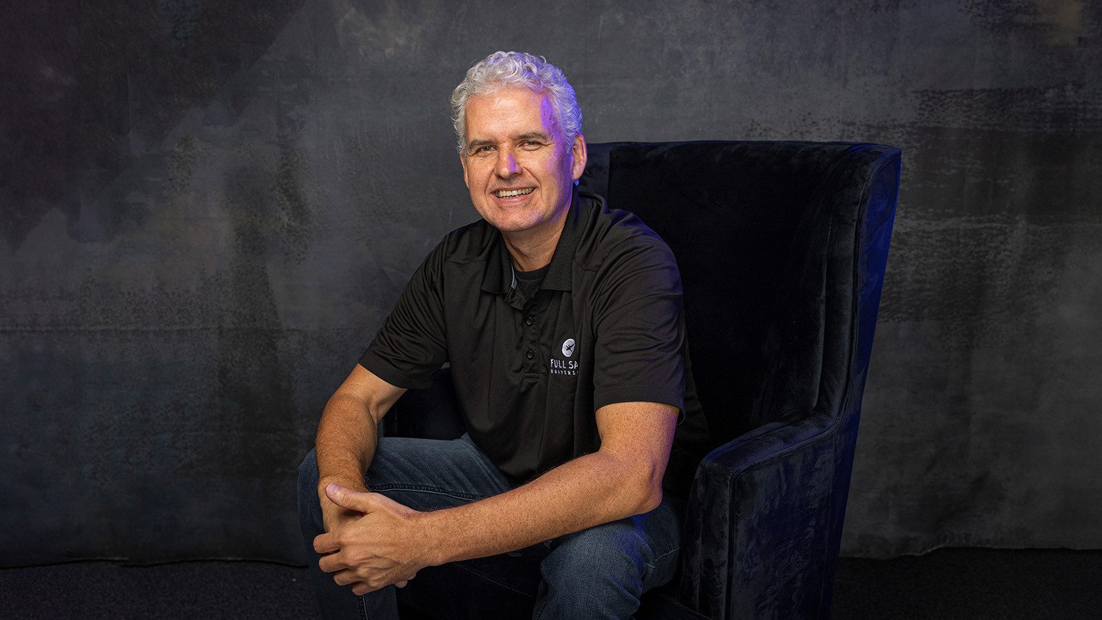 Roy Papp sits in a blue velvet chair in a photography studio. He is smiling and wearing jeans and a black polo shirt with a Full Sail logo.