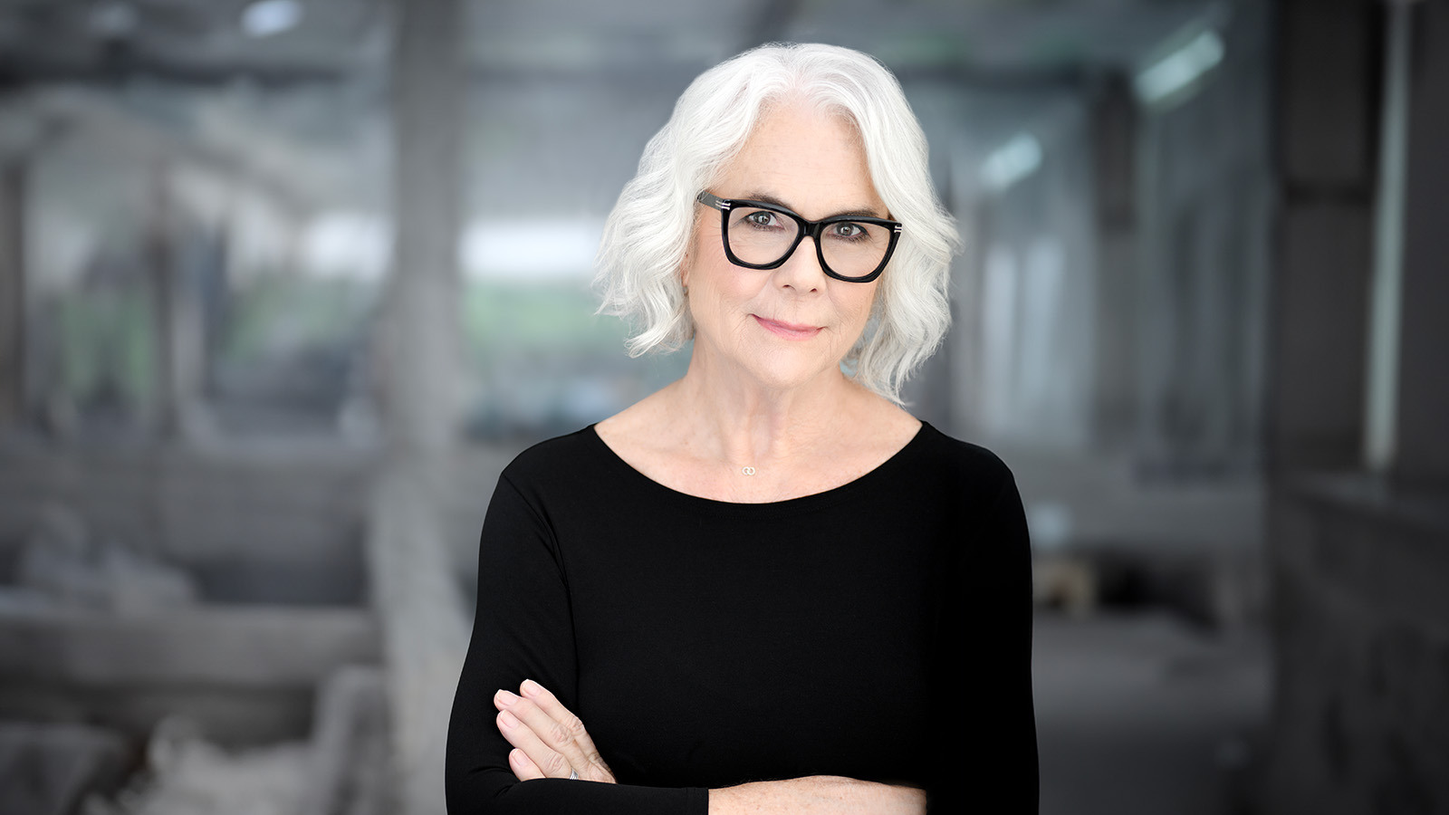 A woman with short white hair and glasses, wearing a black top, smiles confidently with arms crossed against a blurred background.