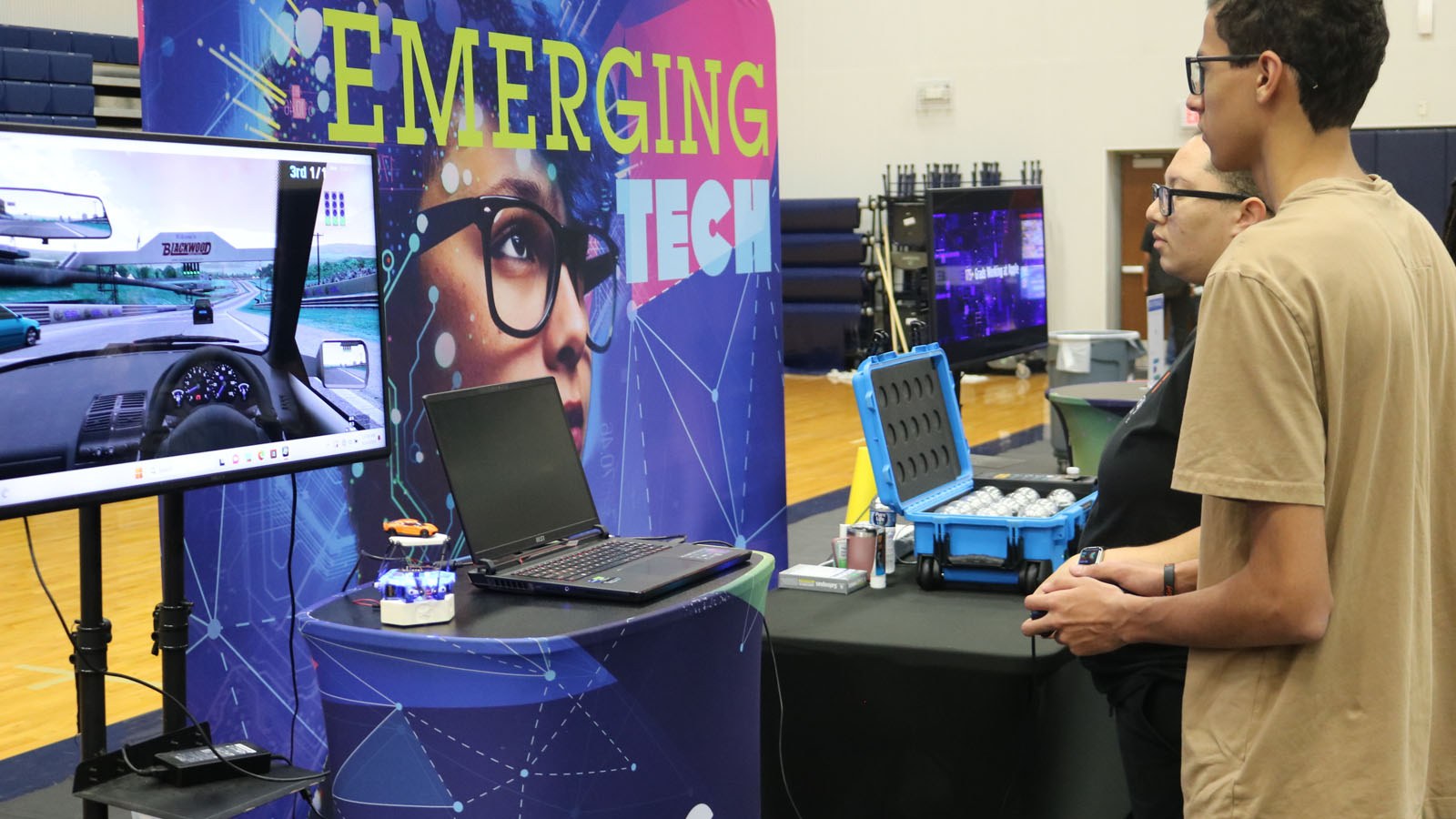 Two people at TechFest explore a driving simulator next to an ‘Emerging Tech’ display with gadgets and a laptop on the table.