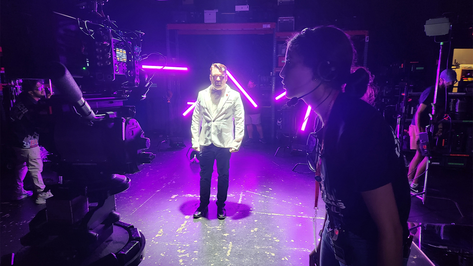 An actor stands in the middle of a set wearing a white suit and bowtie with purple lights around them. A student wears a headset and operates a camera pointed at the actor.