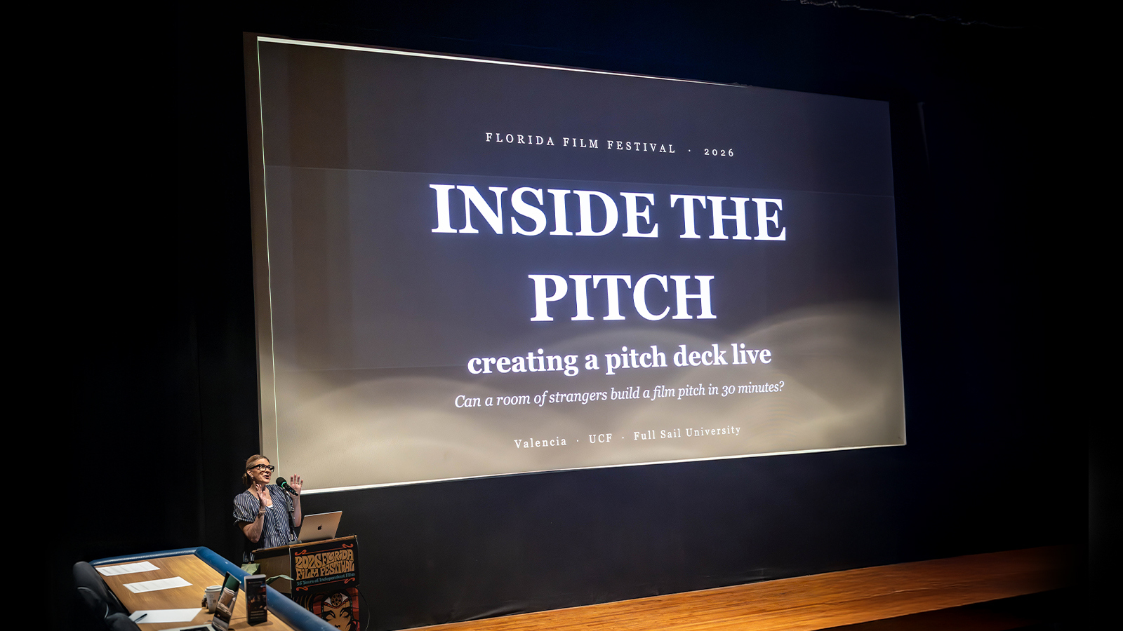 Anne Russell speaks at a podium in front of a large screen displaying “Florida Film Festival 2026 Inside the Pitch: creating a pitch deck live. Can a room of strangers build a film pitch in 30 minutes?”