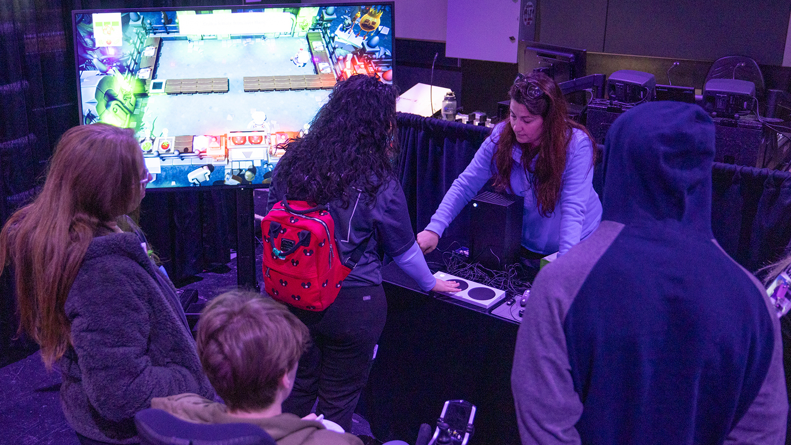 Several people stand around facing a table with adaptive gaming consoles, speakers, and a large screen nearby displaying a game. A staff member stands behind the table.