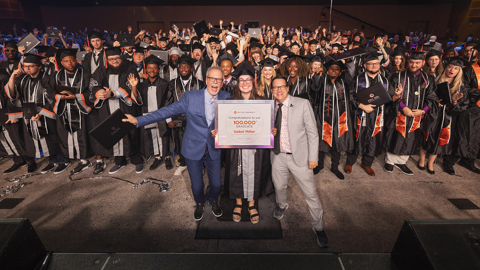 In a large venue full of college graduates, two men in suits stand on opposite sides of a woman in a cap and gown holding a framed photo that reads "Full Sail University - Congratulations to our 100,000th graduate Isabel Miller."