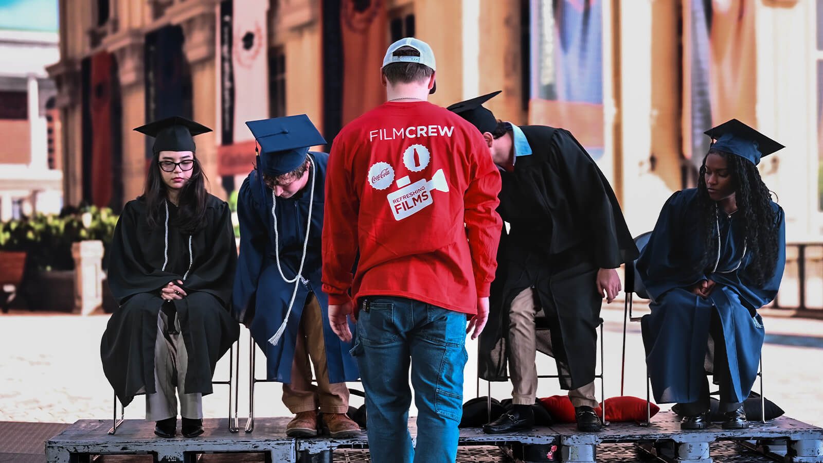 A Coca-Cola Refreshing Films crew member faces seated graduates in caps and gowns during a staged production scene.