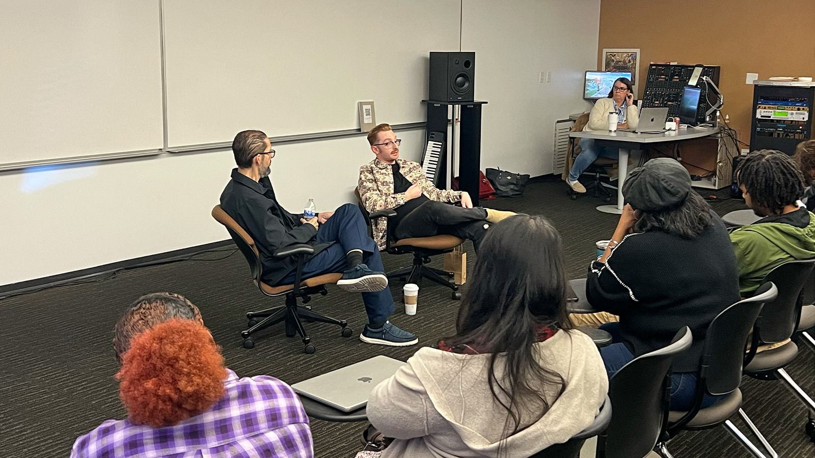 Brad Biwer sits in a chair and speaks to a crowd of students. A staff member sits in a chair next to him and a faculty member sits at their desk behind them.