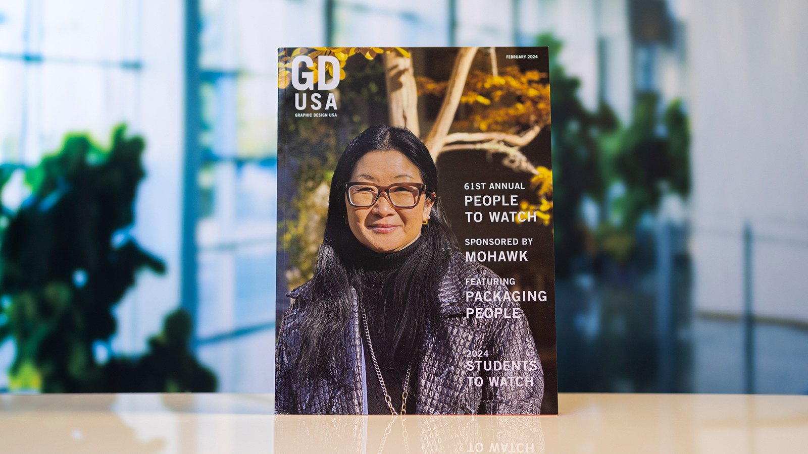 A copy of “Graphic Design USA” magazine depicting a woman wearing glasses and a jacket stands atop a white table.