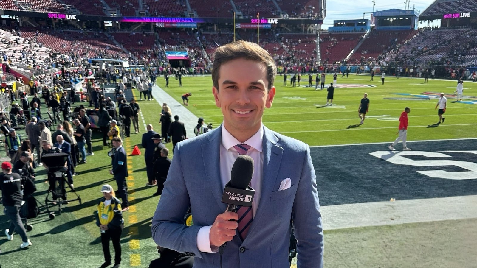 Taylor Schaub stands on the edge of a football field wearing a suit and tie and holding a microphone. Behind him is the football field, stands full of fans, a large screen showing the game, crew on the sidelines, and players on the field.