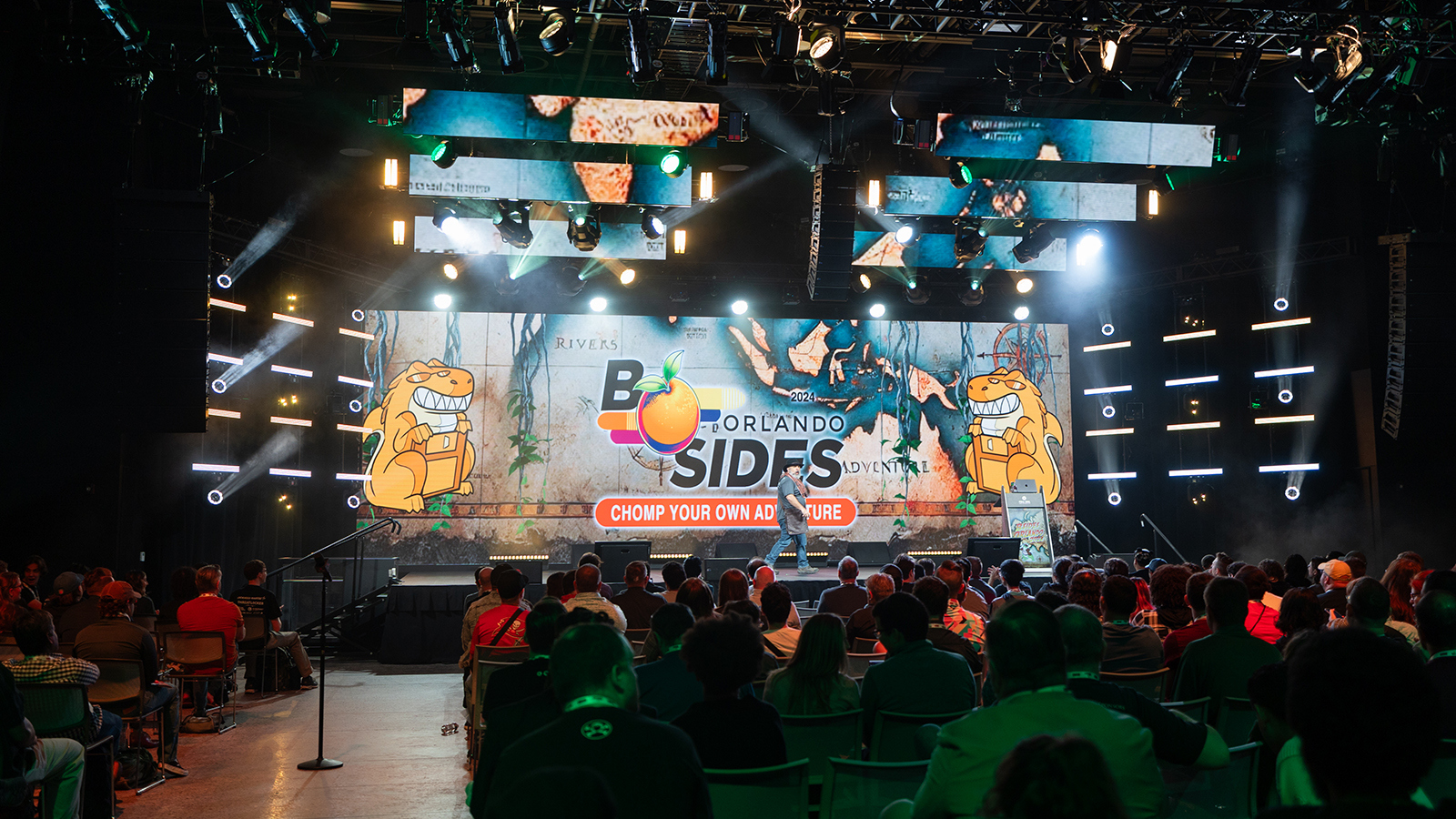A large audience sits in chairs in front of a stage, which has a digital backdrop that says 2024 B-Side Orlando Chomp Your Own Adventure with two cartoon alligators.