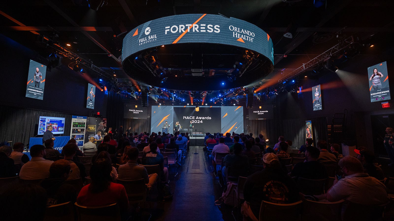 A large audience sits in Full Sail’s esports venue. A stage with a few people standing on it is at the front of the room. The words NACE Awards 2024 are on the wall behind them.
