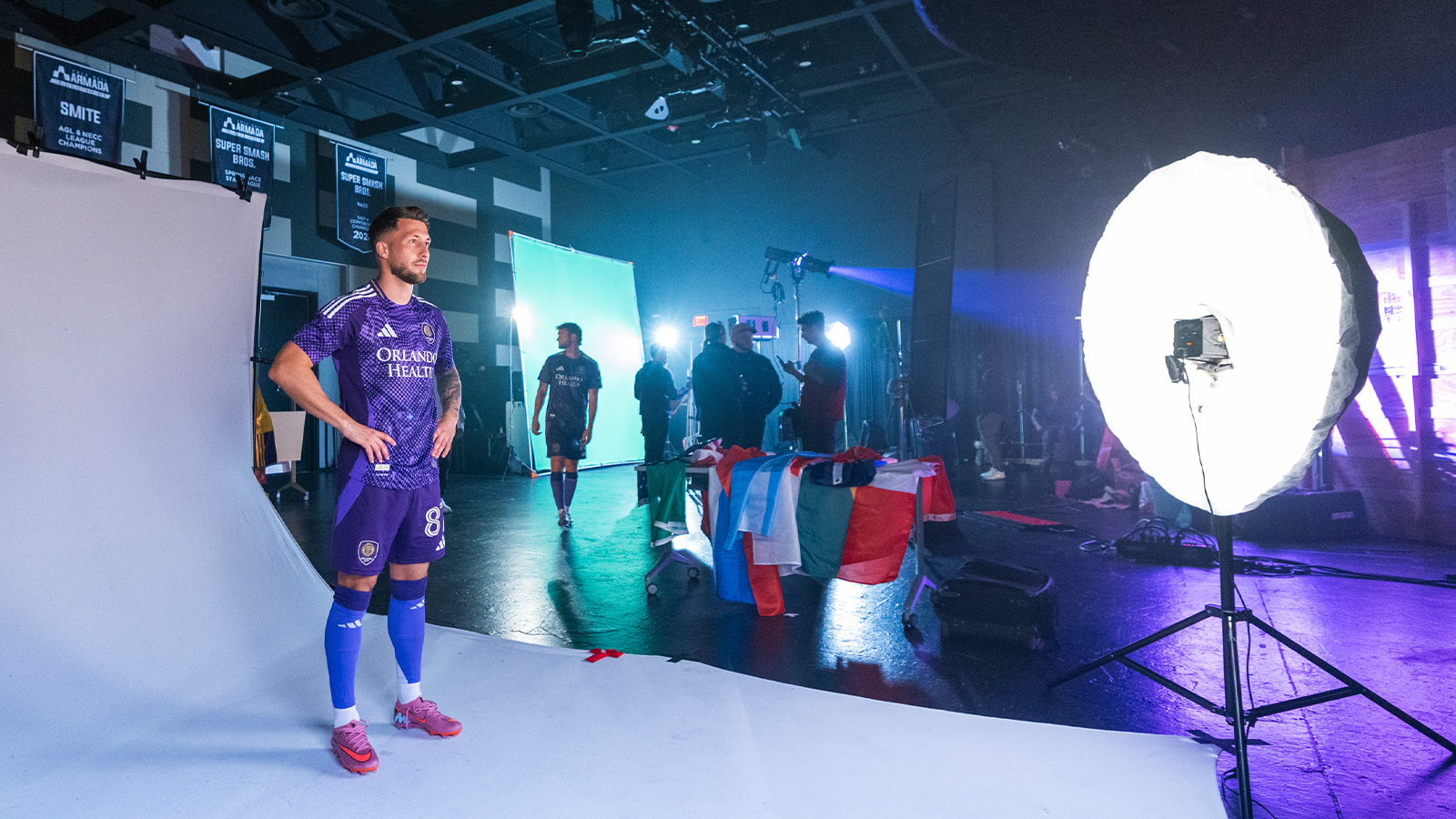 An Orlando City player poses for the camera with their hands on their hips wearing a purple uniform. Production crew members stand around in the background.
