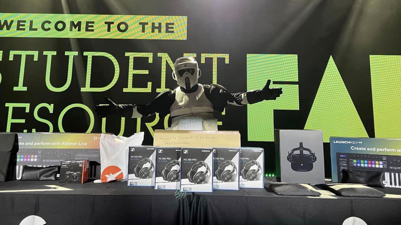 A masked figure stands behind a table piled with boxes of headphones and various technology on stage at the Student Resource Fair.