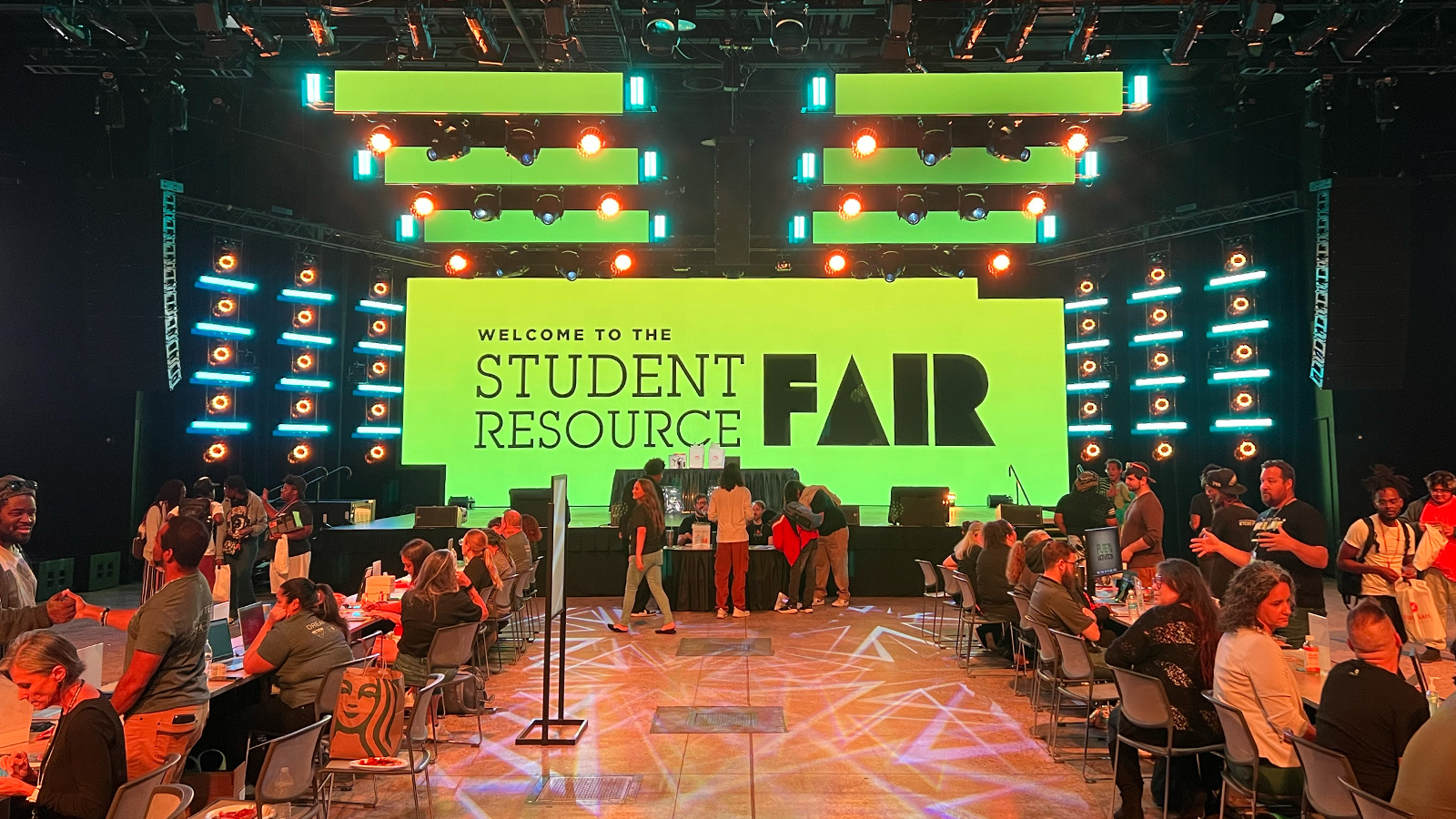 Students and staff sit at tables and interact. A large screen behind them displays “Welcome to the Student Resource Fair” in black text with a green background.