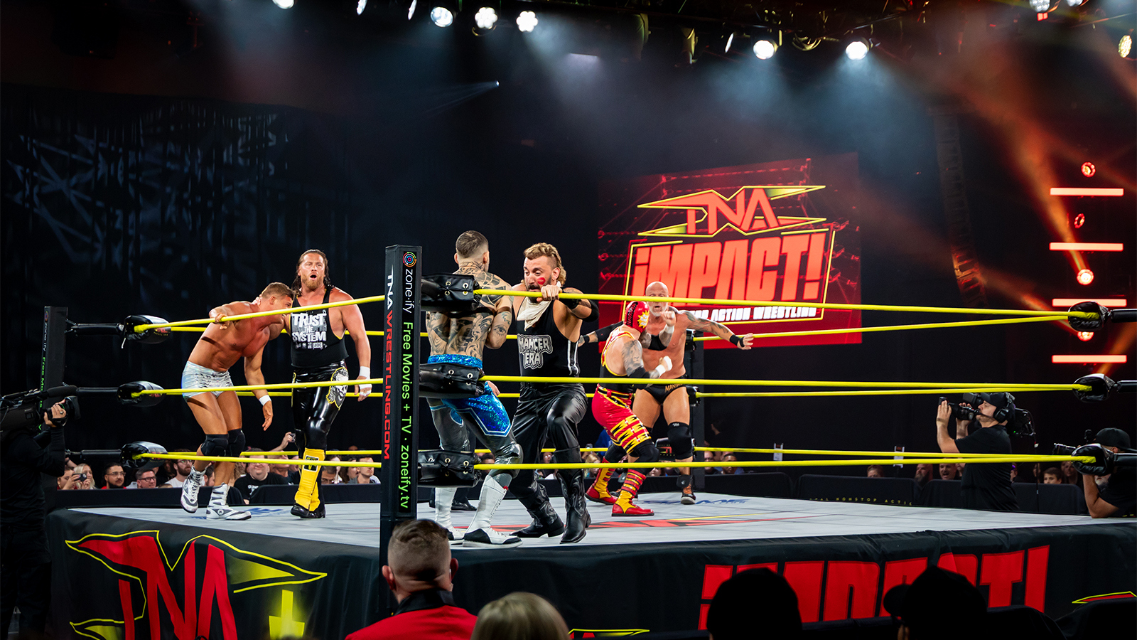 Wrestlers duke it out in the ring during a TNA Impact wrestling match, surrounded by bright stage lights and a cheering crowd in Full Sail's Live Venue.