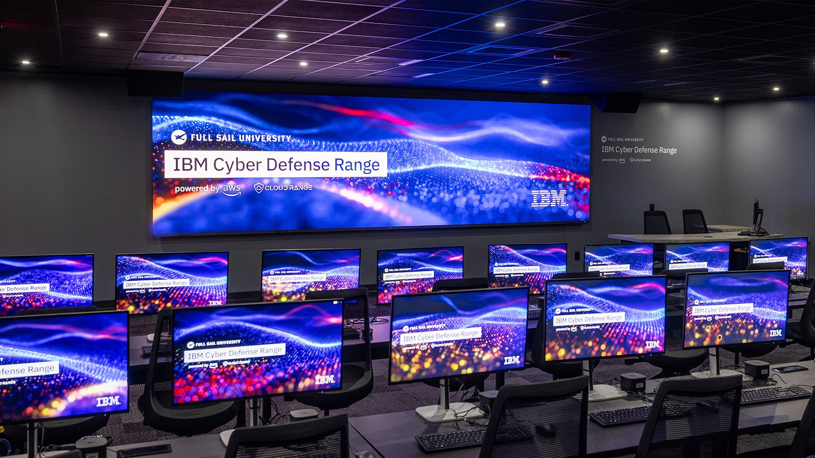 A room with dozens of desktop computers and chairs at long tables. A large screen on the wall reads Full Sail University IBM Cyber Defense Range Powered by AWS and Cloud Range.