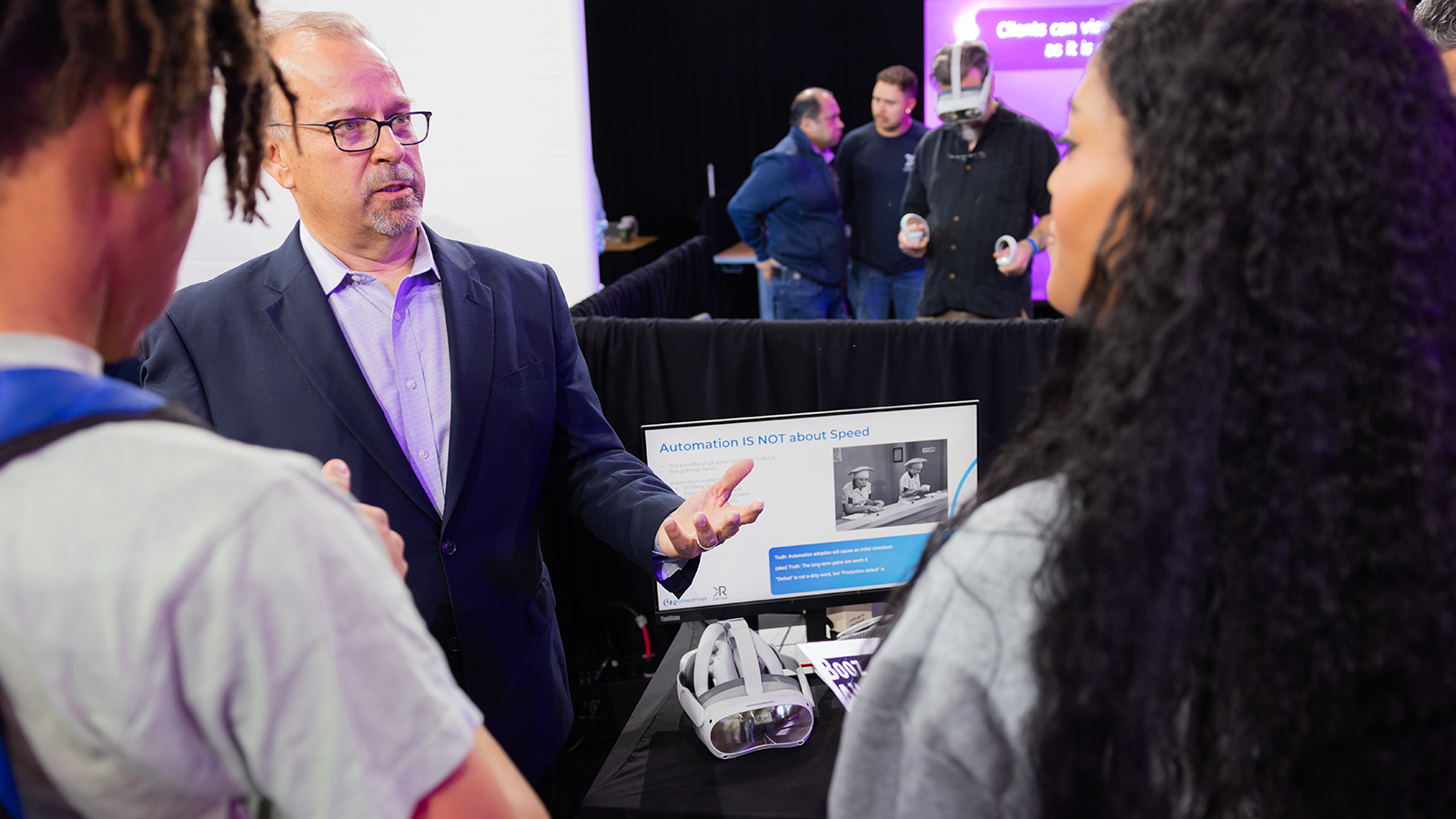 Man wearing glasses and a blue suit speaks to guests at Career Fair while man in back tries on Virtual Reality goggles.
