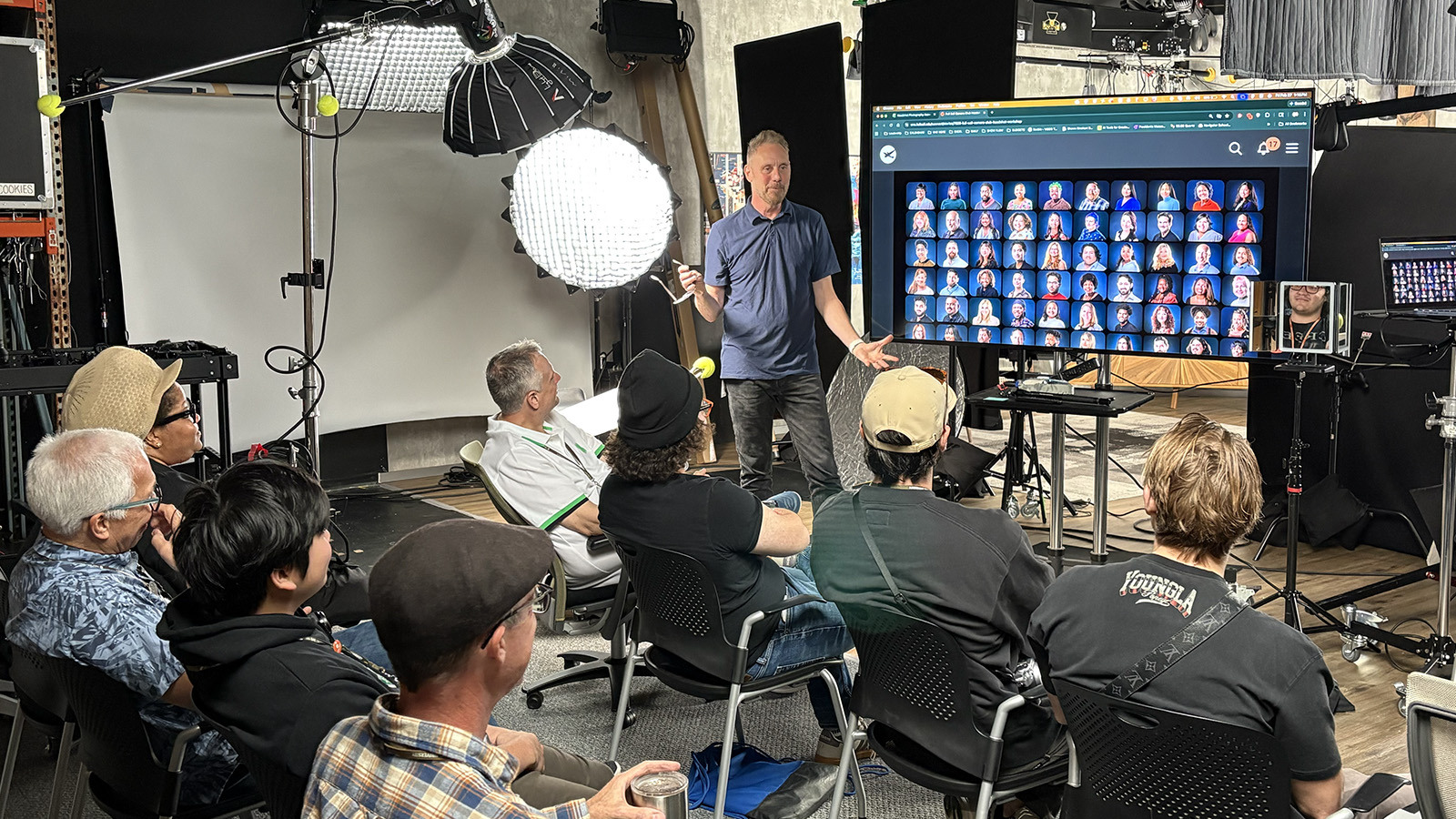 Shawn Rinehart stands in front of an audience of students and faculty next to a screen displaying numerous headshots. Lighting and camera equipment are around the room.