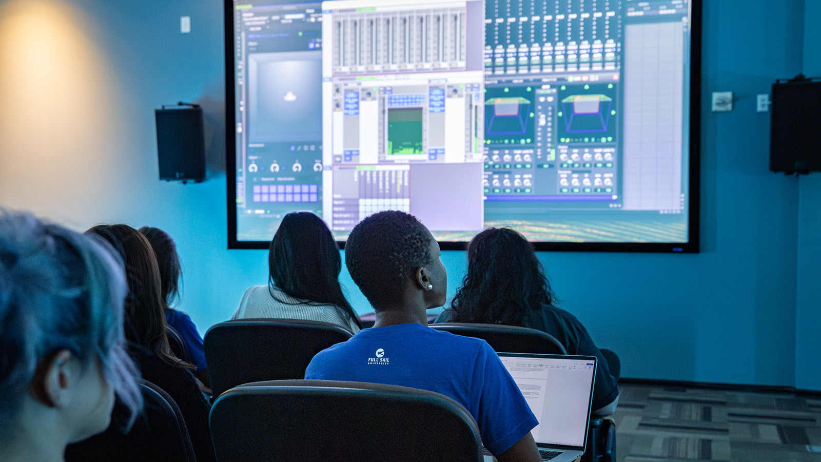 Students sit in a classroom watching a large screen displaying audio mixing software, with laptops open and studio speakers mounted on the walls.