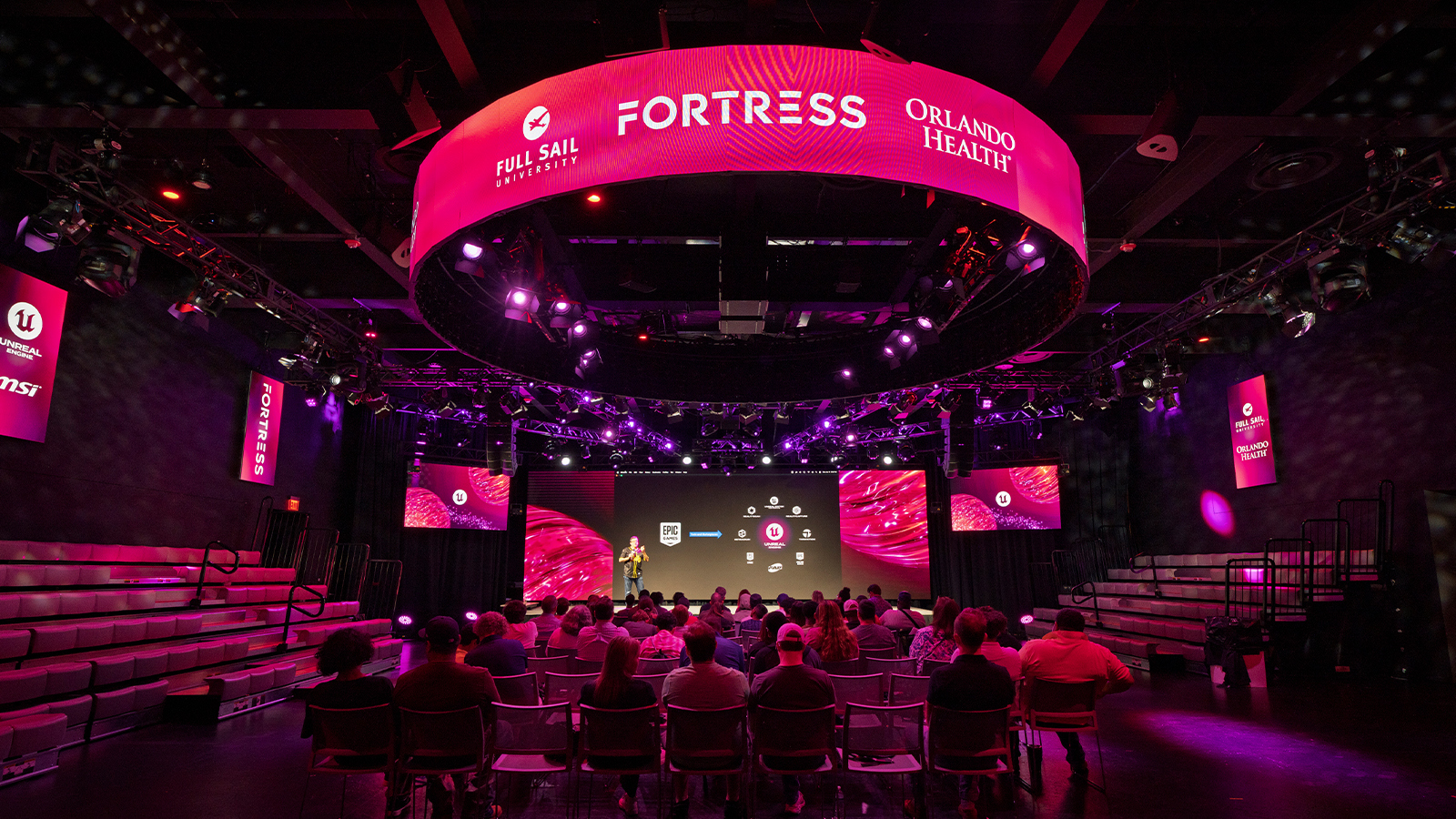 An audience sits in rows of chairs in a large room with a screen and stage. A speaker stands on the stage holding a microphone and red and purple lights shine.