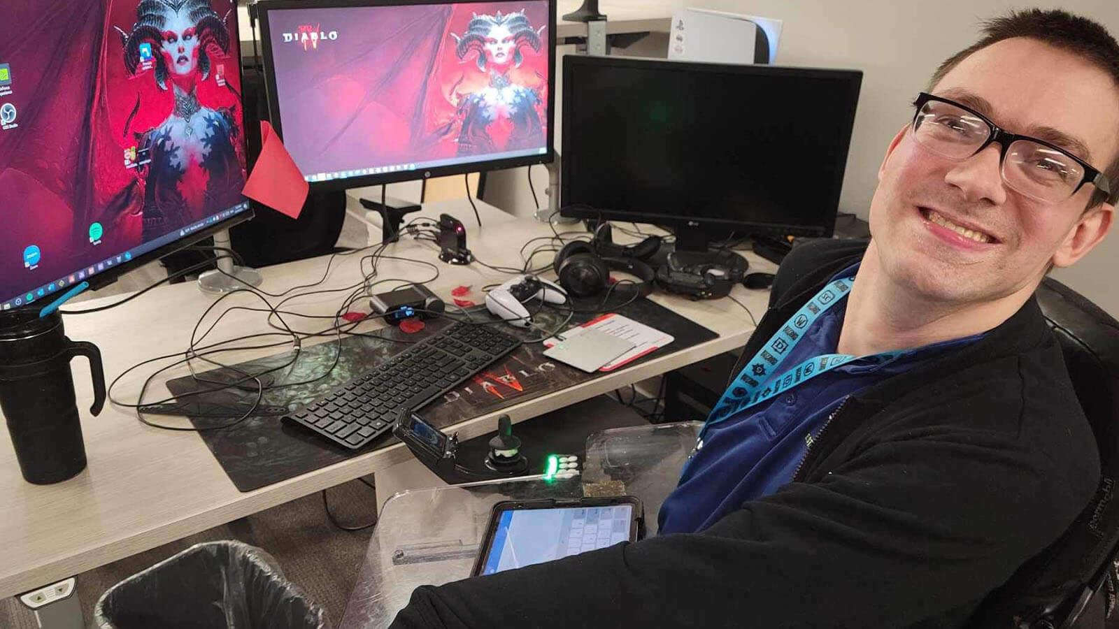 Brock Davis sits at his desk at Blizzard Entertainment. Two computer screens displaying the ‘Diablo IV’ logo are on the desk in front of him.