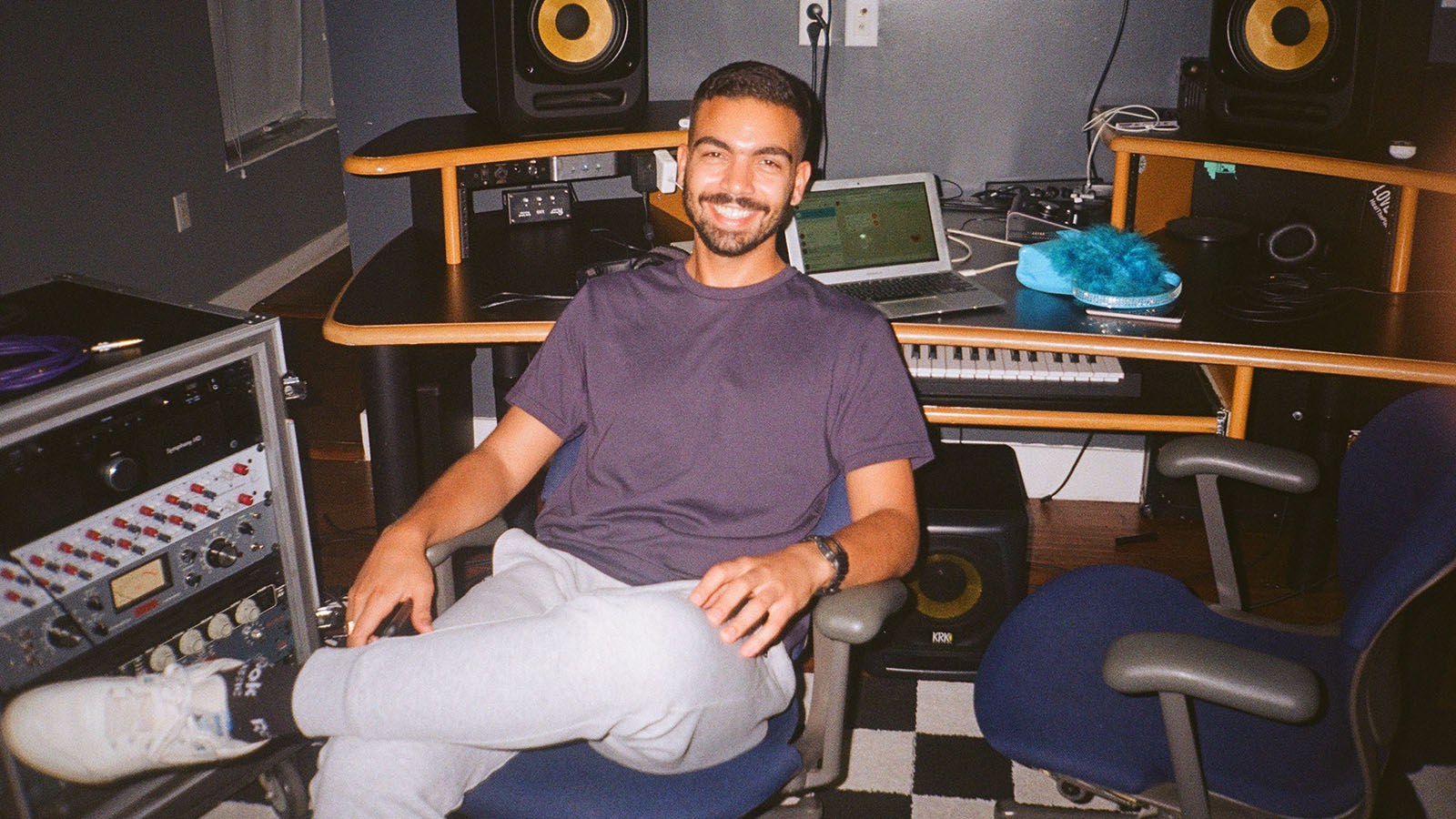 A smiling man is seated casually in a music studio, surrounded by audio equipment, a keyboard, speakers, and a laptop on a desk.