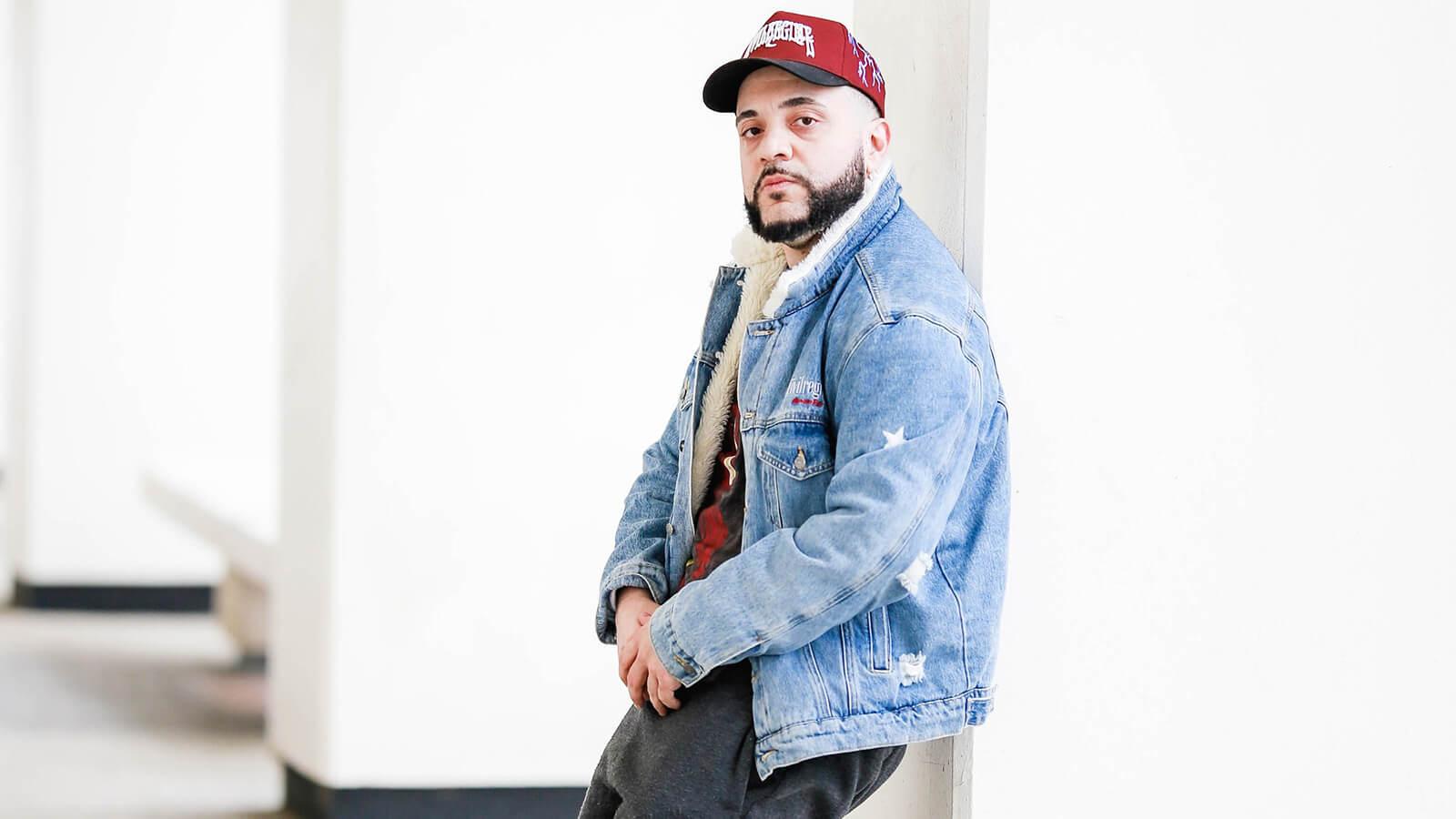 A man wearing a red baseball cap and a denim jacket is pictured leaning against a concrete pillar in a brightly lit room.