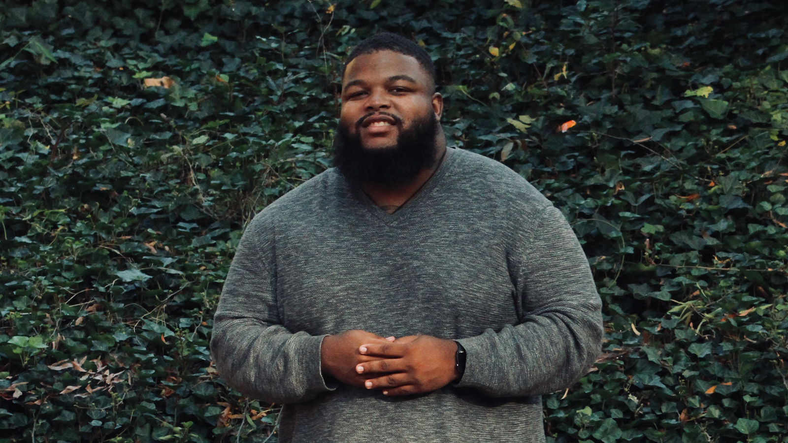 Tariq Ricks poses for the camera in front of a green leaf background. He is wearing a long sleeve grey shirt.