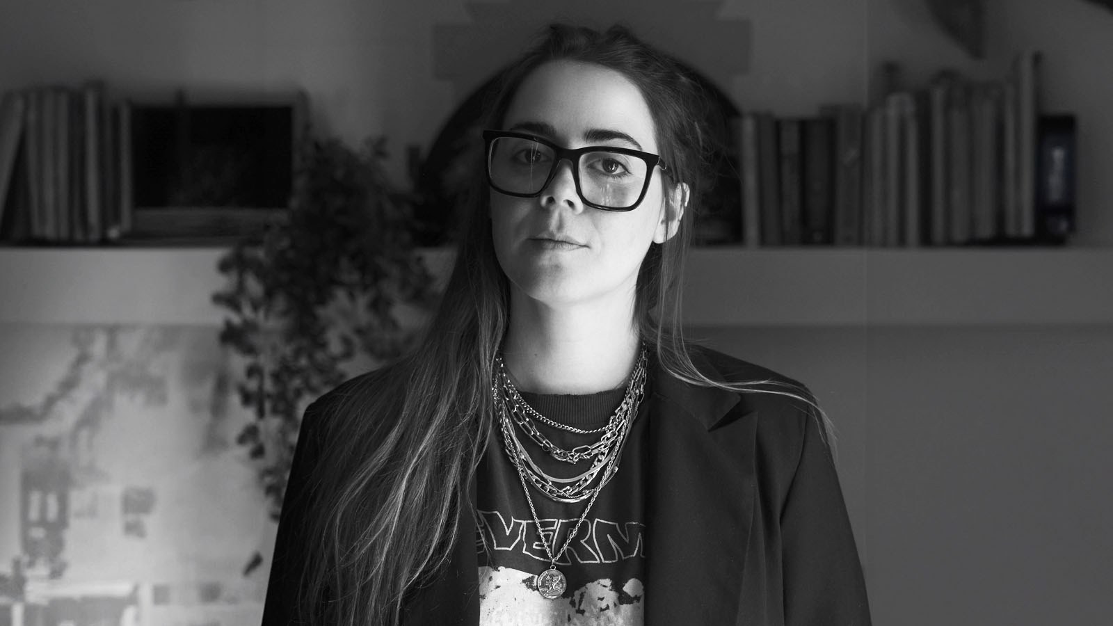 Veronica Concha stands in front of a bookshelf in her home. She is wearing a dark blazer, a band t-shirt, silver necklaces, and glasses.