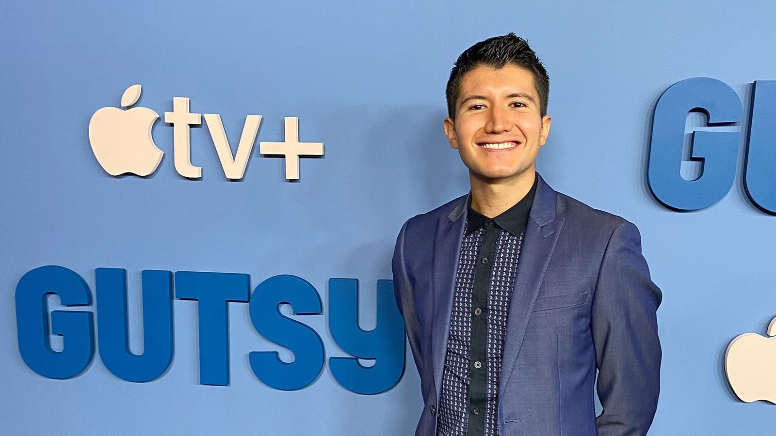 A person with short, dark hair is smiling while wearing a blue suit against a light blue step and repeat that shows the logos for ‘Gutsy’ and Apple TV+.