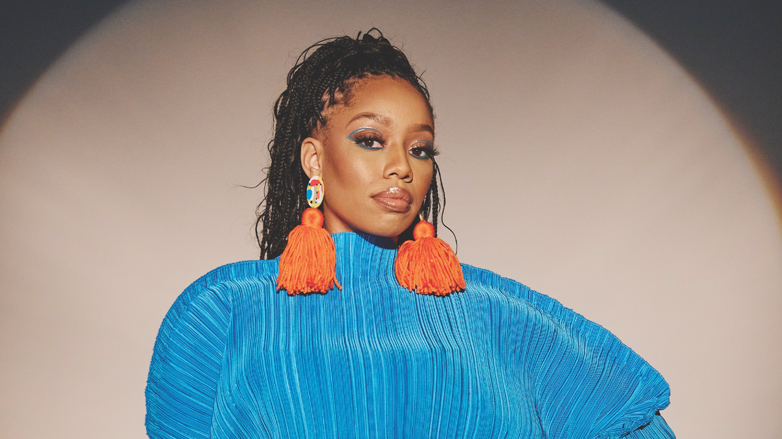 DOE stands against a white wall. She has long braids tied up in a ponytail and wears a bright blue sweater and earrings with orange tassels.