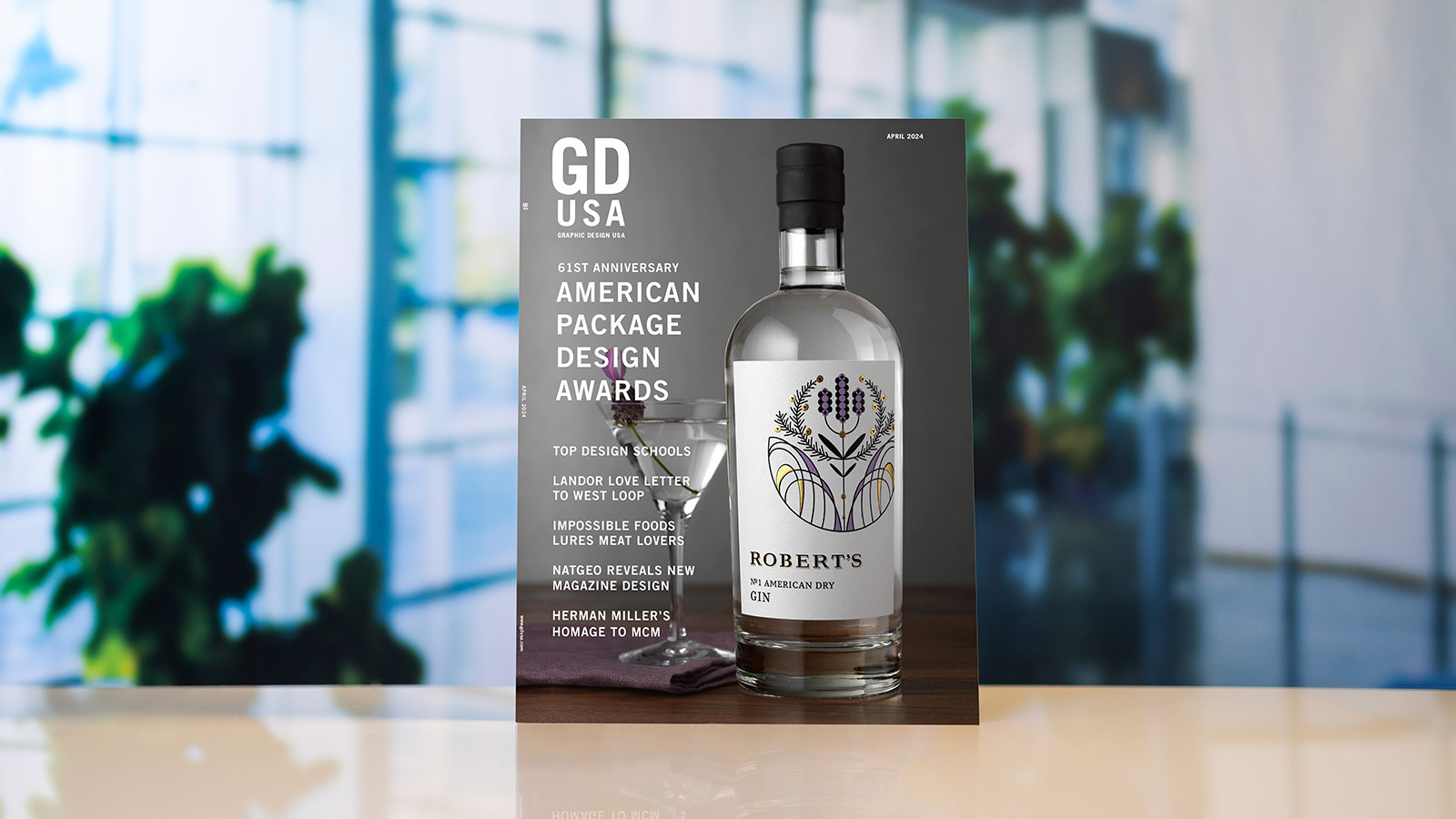 A copy of 'Graphic Design USA' magazine sits atop a white surface against a blurred background. The magazine cover depicts a bottle of gin and a martini glass against a gray background and reads '61st Anniversary American Package Design Awards.