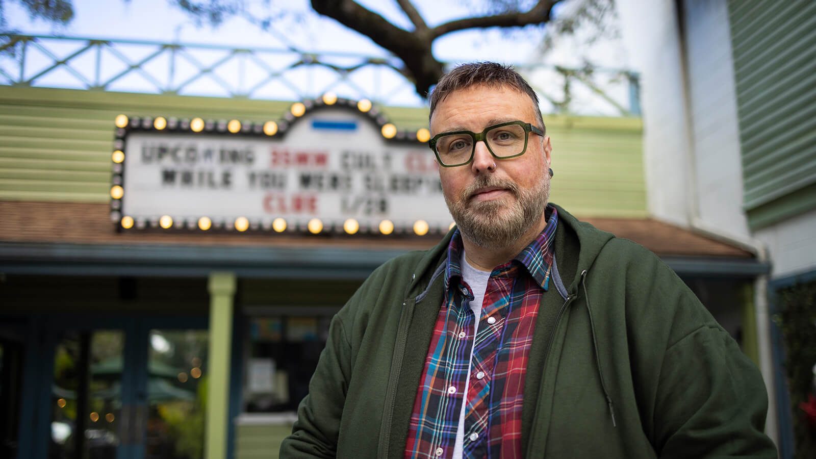 Aaron B. Koontz stands in front of a movie theater. He is wearing green glasses, a green sweatshirt, and a collared plaid shirt.