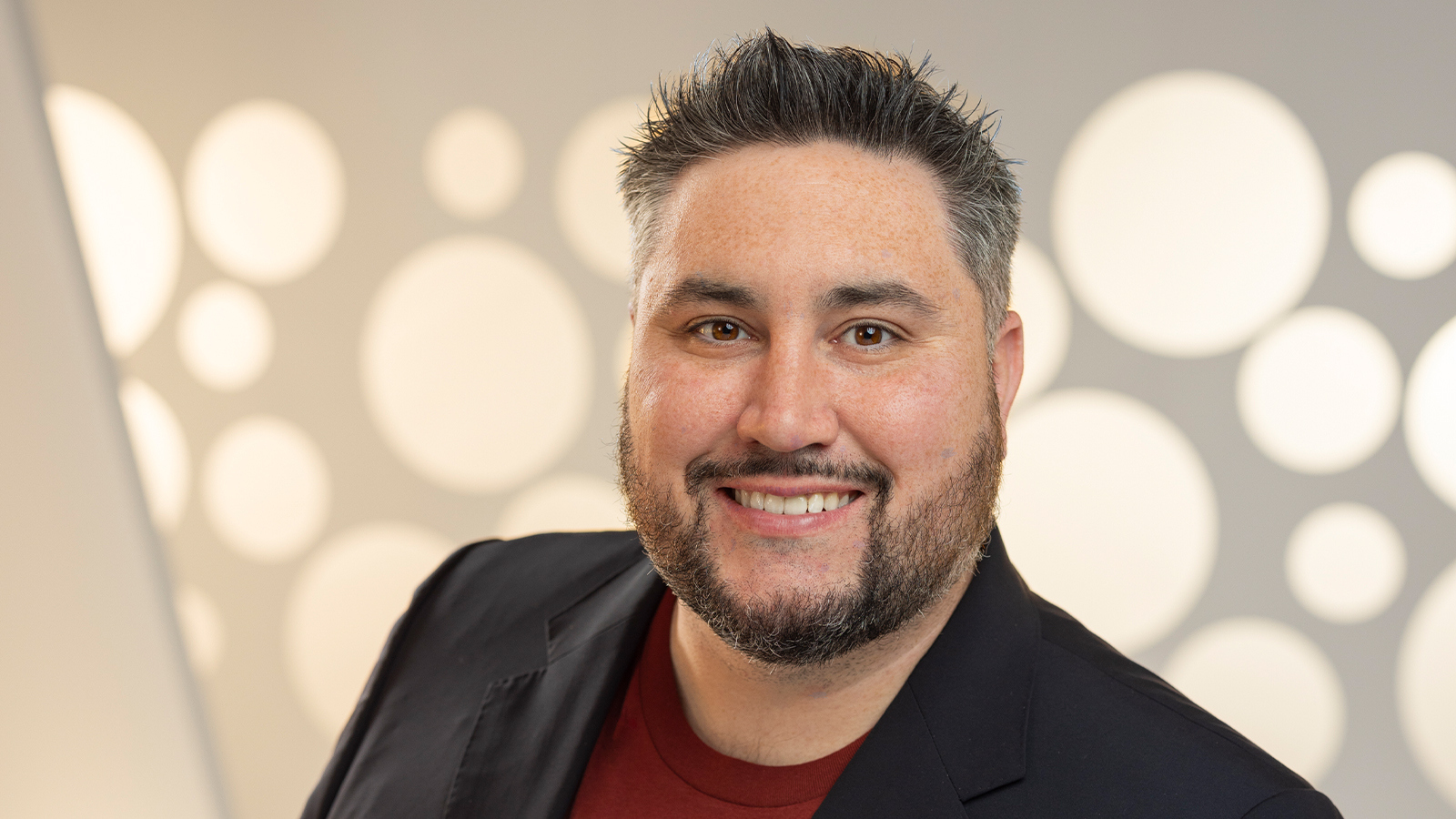 Michael La Plante smiles at the camera. He is wearing a red t-shirt with a black blazer and standing in front of a white backdrop.