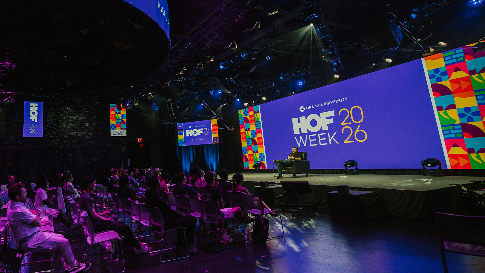 A speakers sits in a chair on a stage with a large screen behind them displaying “HOF Week 2026” and colorful graphics. An audience sits in rows of chairs facing the stage.