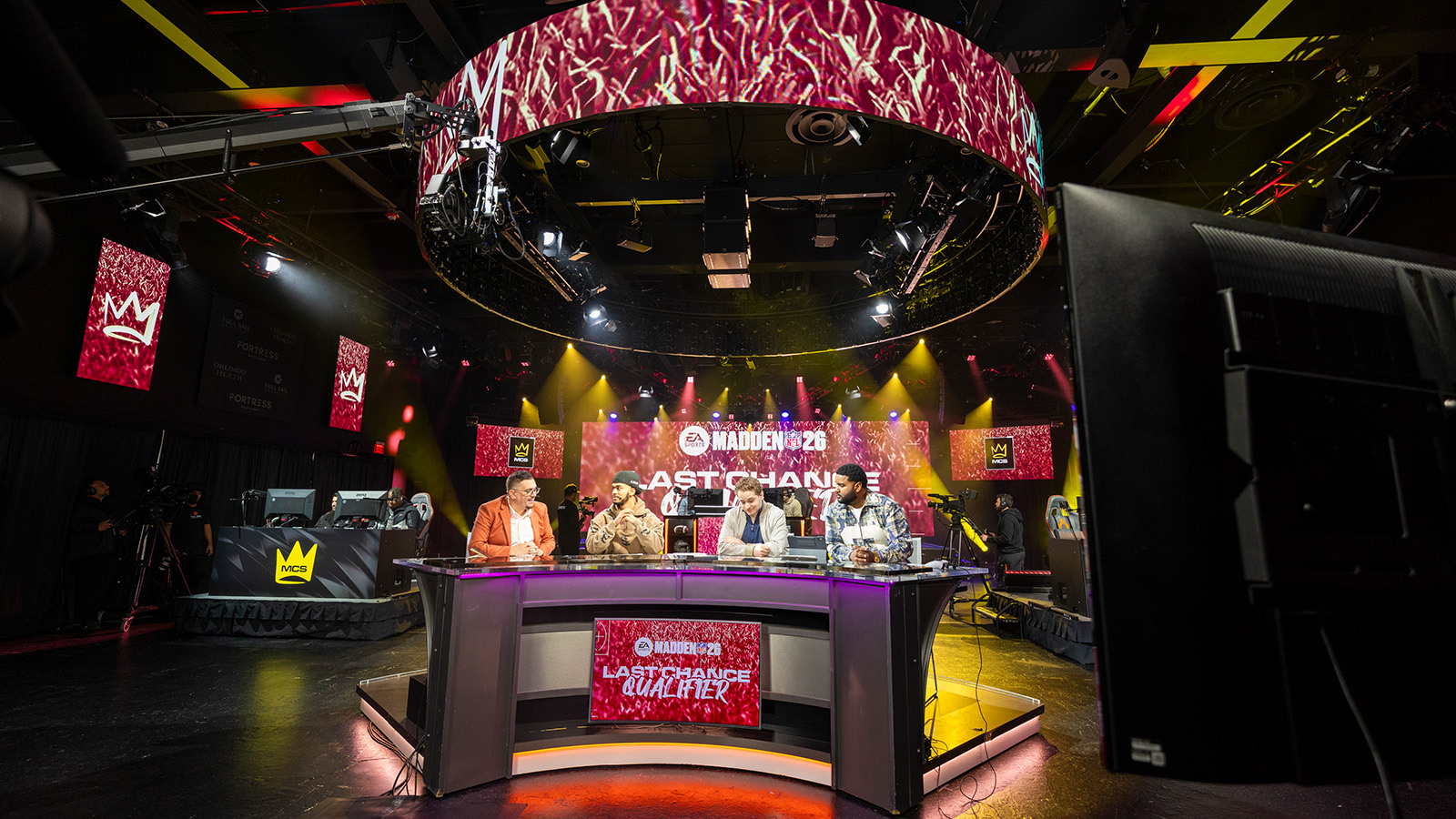 Analysts sit at a broadcast desk beneath a glowing “Madden NFL 26” ring as cameras roll on a live esports studio set.