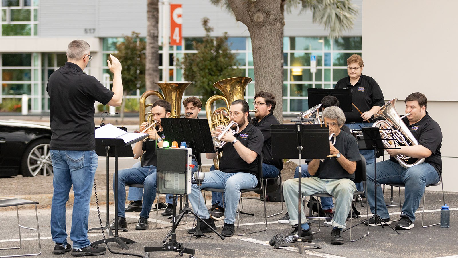 Members of the Brass Band of Central Florida perform in a parking lot, sitting down and reading from their music stands while a conductor stands at the front.